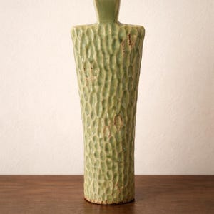 May include: A textured, light green ceramic vase with a unique shape. The vase has a rectangular top and a cylindrical body with a rough, dimpled surface. The vase is approximately 10 inches tall.