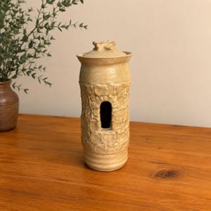 May include: A cylindrical ceramic vessel with a light tan, matte finish. The exterior features a textured, hand-sculpted stone wall pattern with a single rectangular opening in the center. It includes a matching lid with a small, raised knob.