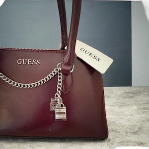 May include: A burgundy Guess handbag with silver-tone chain detailing and a decorative lock charm. The bag features a top handle and a zipper closure. A Guess tag is attached to the handle.