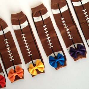 May include: Six pairs of brown and white football-themed knee-high socks with colorful bows attached to the top of each sock.