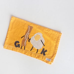 Puede incluir: Toalla de cocina amarilla con un dise&ntilde;o bordado de un figur&iacute;n de palo que se da la mano con un huevo frito. El figur&iacute;n de palo sostiene una letra G y el huevo sostiene una letra K.