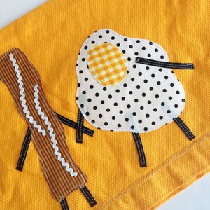 Puede incluir: Una toalla de cocina amarilla con un dise&ntilde;o bordado de un huevo frito con lunares negros y una yema amarilla a cuadros, y dos palitos marrones y blancos.