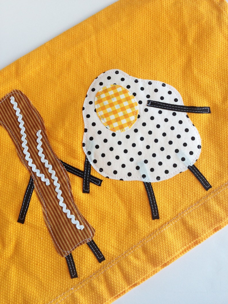 Puede incluir: Una toalla de cocina amarilla con un dise&ntilde;o bordado de un huevo frito con lunares negros y una yema amarilla a cuadros, y dos palitos marrones y blancos.