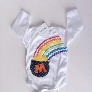 May include: White baby onesie with a rainbow and a pot of gold with the letter M on it. The onesie has the text "kakabaka" and "www.kakabaka.com" on the tag.
