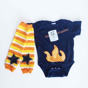 May include: A navy blue baby onesie with a felt applique of a campfire and a marshmallow. The onesie has a white, orange, and yellow striped leg warmers with black felt stars.