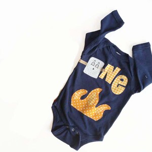 May include: A navy blue long-sleeve onesie with a yellow and white appliqué of a campfire and the word "one" in yellow and white gingham.