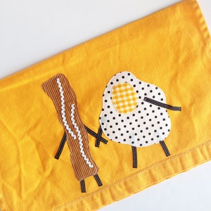 May include: A yellow kitchen towel with an embroidered design of a fried egg and a stick of butter. The egg has a yellow and white checkered yolk and black polka dots.