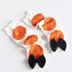 Puede incluir: Un par de calcetines blancos hasta la rodilla con dise&ntilde;os de baloncesto naranja y lazos negros.