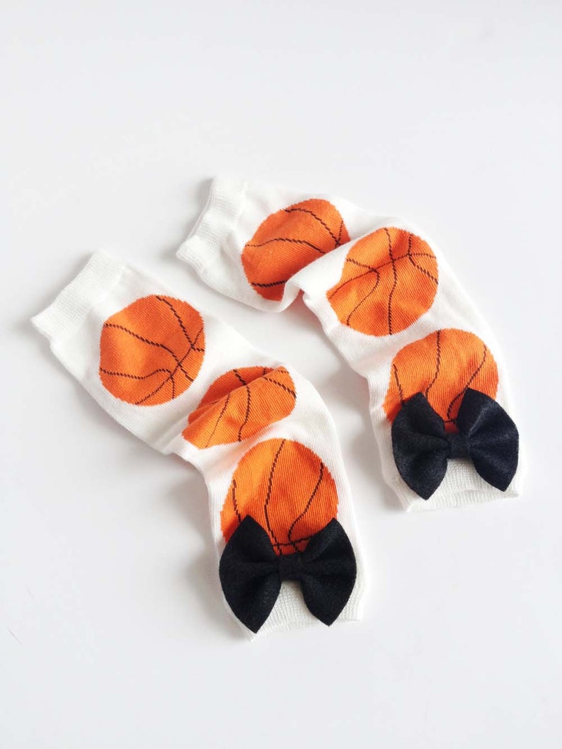 Puede incluir: Un par de calcetines blancos hasta la rodilla con dise&ntilde;os de baloncesto naranja y lazos negros.
