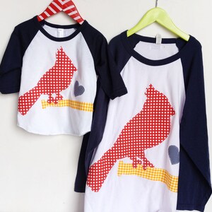 May include: Two white t-shirts with red and white polka dot cardinal appliques. The shirts have navy blue sleeves and are on hangers.