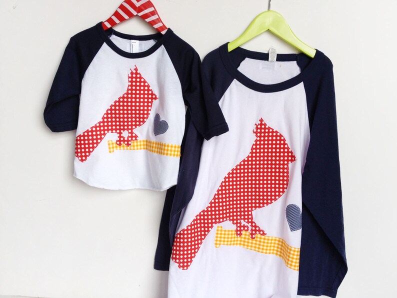 May include: Two white t-shirts with red and white polka dot cardinal appliques. The shirts have navy blue sleeves and are on hangers.