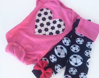 May include: A pink baby bodysuit with a soccer ball heart design, paired with black and white soccer ball patterned leg warmers. The leg warmers have red bows at the ankles. The outfit is designed for a baby.
