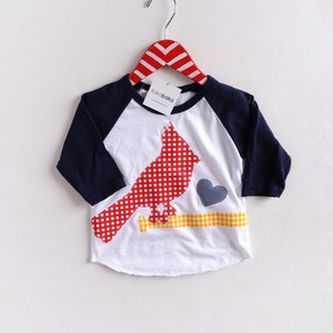 May include: White baseball tee with a red cardinal applique with white polka dots, a blue heart applique with white polka dots, and a yellow branch applique with white and yellow checkered pattern. The shirt has navy blue sleeves.