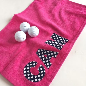 May include: Pink golf towel with a navy blue and white polka dot "CAM" embroidered on it. Three white golf balls are on the towel.