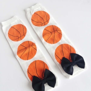 Puede incluir: Un par de polainas blancas con dise&ntilde;os de baloncesto naranja y lazos negros.