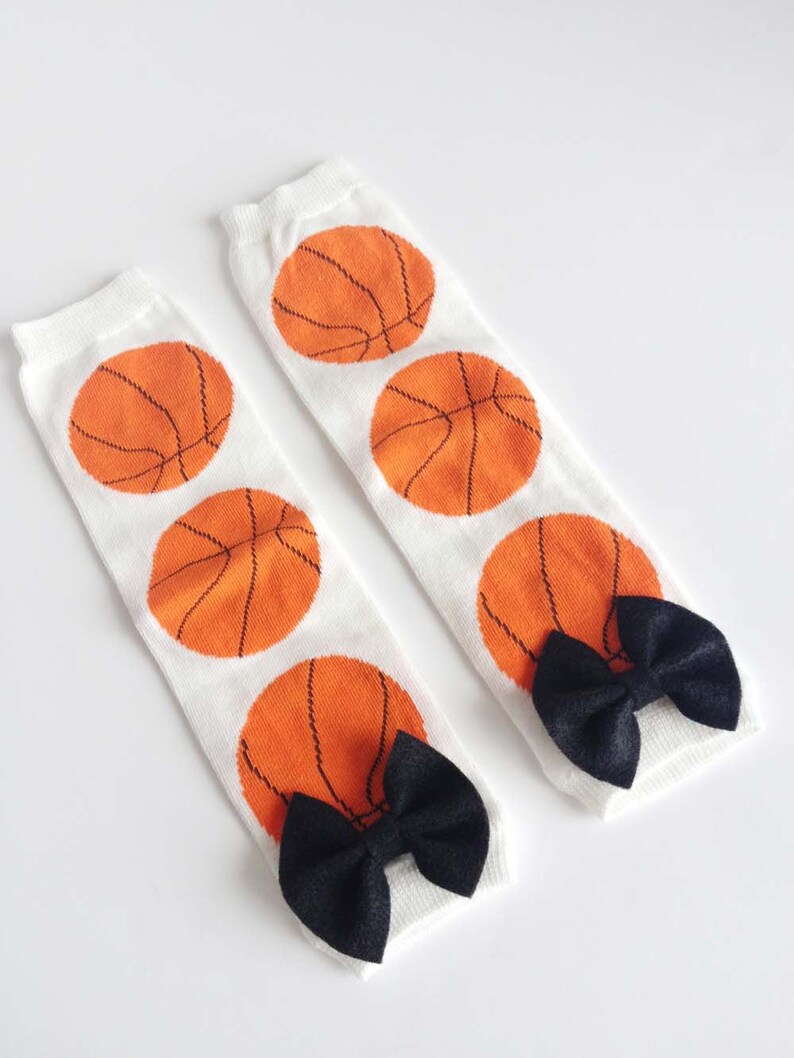 Puede incluir: Un par de polainas blancas con dise&ntilde;os de baloncesto naranja y lazos negros.
