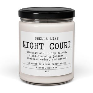 May include: A glass jar candle with a black lid and white wax. The label reads: SMELLS LIKE NIGHT COURT, sea-salt air, crisp citrus, night-blooming jasmine, shadowed cedar, and dreams, 50 HOURS OF NIGHT COURT FLAME, NATURAL SOY WAX, 9OZ.