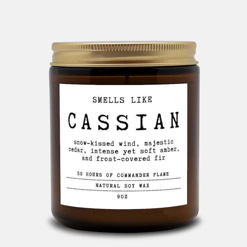 May include: A brown glass candle jar with a gold metal lid and a white label. The label reads: SMELLS LIKE CASSIAN, snow-kissed wind, majestic cedar, intense yet soft amber, and frost-covered fir, 50 HOURS OF COMMANDER FLAME, NATURAL SOY WAX, 9OZ.