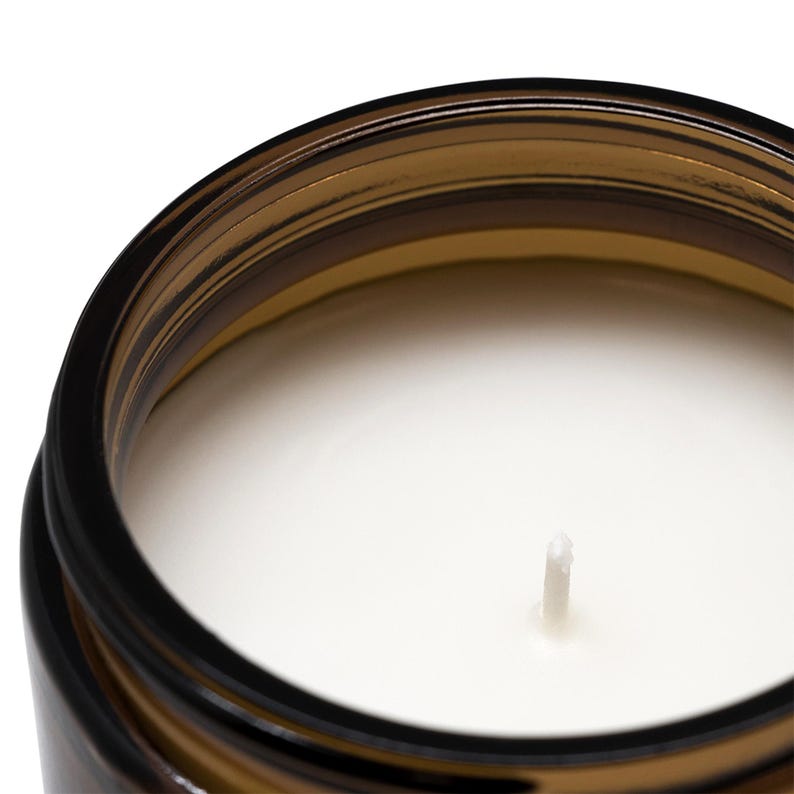May include: A top-down view of a cylindrical amber glass jar containing a smooth, solid white candle wax. A single, centered white cotton wick protrudes from the surface of the wax. The jar features a thick, dark brown glass rim with a threaded top.