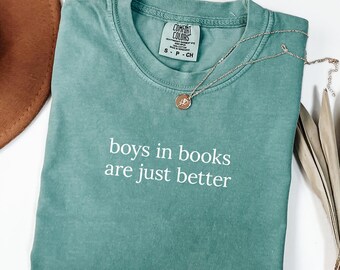 May include: A teal Comfort Colors t-shirt with the phrase "boys in books are just better" in white. A gold-colored necklace with a small pendant rests on the shirt. A brown hat and dried leaves are in the background.