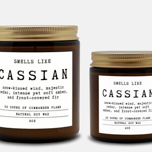 May include: Two amber glass candles with gold lids and white labels. The labels read: SMELLS LIKE CASSIAN, snow-kissed wind, majestic cedar, intense yet soft amber, and frost-covered fir. The larger candle is 9oz, the smaller is 4oz, both soy wax.