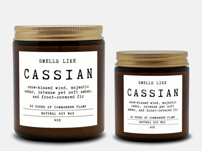 May include: Two amber glass candles with gold lids and white labels. The labels read: SMELLS LIKE CASSIAN, snow-kissed wind, majestic cedar, intense yet soft amber, and frost-covered fir. The larger candle is 9oz, the smaller is 4oz, both soy wax.