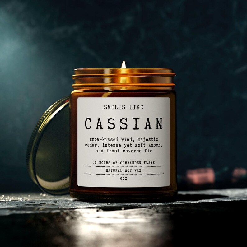 May include: A lit candle in an amber glass jar with a gold lid. The white label reads: SMELLS LIKE CASSIAN, snow-kissed wind, majestic cedar, intense yet soft amber, and frost-covered fir, 50 HOURS OF COMMANDER FLAME, NATURAL SOY WAX, 9OZ.