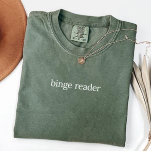 May include: An olive green t-shirt with the words "binge reader" in white. A gold-colored necklace with a pendant is draped across the shirt. The shirt is folded, and a brown hat is partially visible.