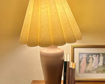 May include: A vintage table lamp with a pleated, light yellow lampshade. The base is a soft pink colour. Several books and a framed picture are next to the lamp on a wooden surface. The background is a light yellow wall.