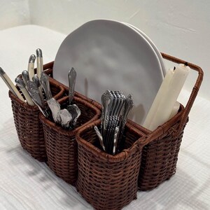 Puede incluir: Un portacubiertos de mimbre tejido marrón con múltiples compartimentos. El soporte contiene cubiertos, platos blancos y velas. El asa es arqueada. El soporte está sobre una superficie blanca.