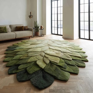 Puede incluir: Una alfombra grande y redonda con diseño de hoja, en tonos verdes. La alfombra está colocada sobre un suelo de madera, con un sofá y ventanas al fondo. El diseño de la alfombra se asemeja a una gran hoja o pétalo.
