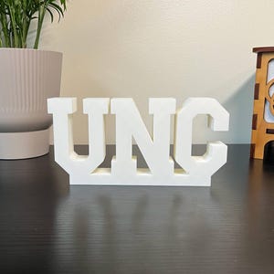May include: A white, three-dimensional sign spells out "UNC" in bold, block letters. The letters are connected at the base, creating a solid structure. The sign is displayed on a dark surface.