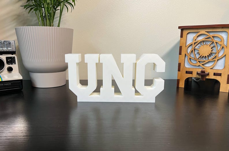 May include: A white, three-dimensional sign spells out "UNC" in bold, block letters. The letters are connected at the base, creating a solid structure. The sign is displayed on a dark surface.