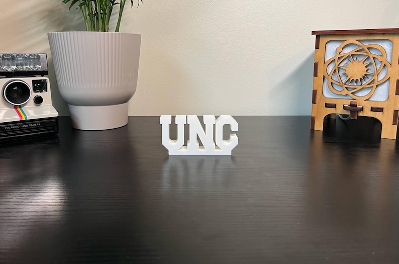 May include: A white, block-lettered "UNC" sign stands on a dark surface. A vintage Polaroid camera with a rainbow stripe and a large white planter with a plant are on the left. A wooden decorative piece is on the right.