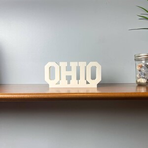 May include: A cream-colored wooden sign displaying the word "OHIO" in a bold, block letter font. The sign is placed on a wooden shelf, alongside a framed picture, a glass jar with decorative elements, and a potted plant.