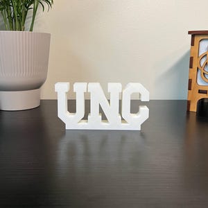 May include: A white, three-dimensional sign that spells out "UNC" in bold, block letters. The letters are evenly spaced and stand upright on a dark surface. The sign is a decorative item.