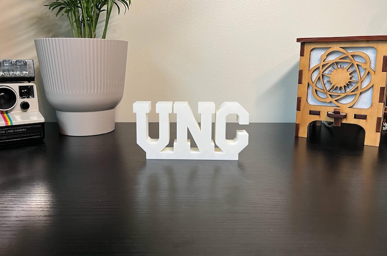 May include: A white, three-dimensional sign that spells out "UNC" in bold, block letters. The letters are evenly spaced and stand upright on a dark surface. The sign is a decorative item.