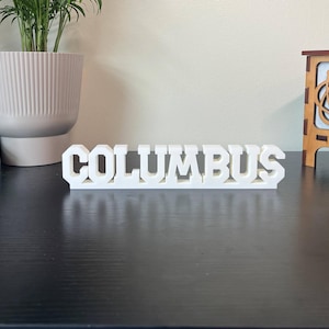 May include: A white ceramic mug with a black handle and the text "But First, Coffee" in a black, bold font. The mug is sitting on a wooden surface. The mug is approximately 4 inches tall.