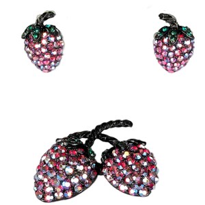 May include: A jewelry set consisting of two strawberry-shaped stud earrings and a matching brooch featuring two strawberries. Each piece is encrusted with small, iridescent pink and red crystals, accented with dark metal stems and green leaves.