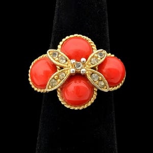 May include: A gold-toned ring featuring a flower design with four red, round stones and a central butterfly-shaped accent. The butterfly has clear, sparkling stones. The ring is displayed on a black velvet ring holder.