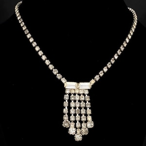 May include: A silver-toned necklace with a series of small, round, clear gemstones forming the chain. The pendant features a rectangular bar with dangling rows of square and round gemstones, creating a tiered effect. The necklace is displayed on a black mannequin.