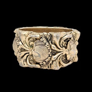 May include: Ornate gold-colored ring with detailed floral and scrollwork patterns. The ring has a wide band with a textured surface and a vintage aesthetic. The design features symmetrical elements and a polished finish.