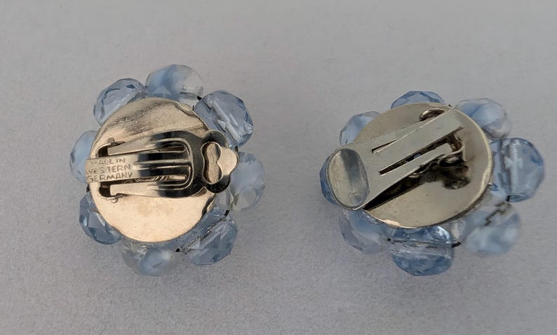May include: A pair of vintage clip-on earrings. Each earring features a cluster of light blue faceted beads surrounding a silver-tone metal backing. The clip mechanism is visible, and the earrings are marked "MADE IN WESTERN GERMANY".