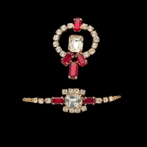 May include: A gold-toned jewelry set featuring a necklace pendant and a bracelet. The pendant is a circle of clear crystals with a central rectangular clear stone and red rectangular stones. The bracelet has a similar design.