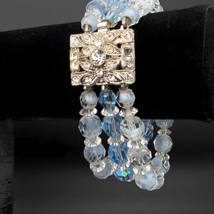 May include: A vintage bracelet featuring three strands of light blue and clear crystal beads. The bracelet has a silver-tone square clasp adorned with clear rhinestones and intricate floral designs. The beads are faceted, reflecting light.