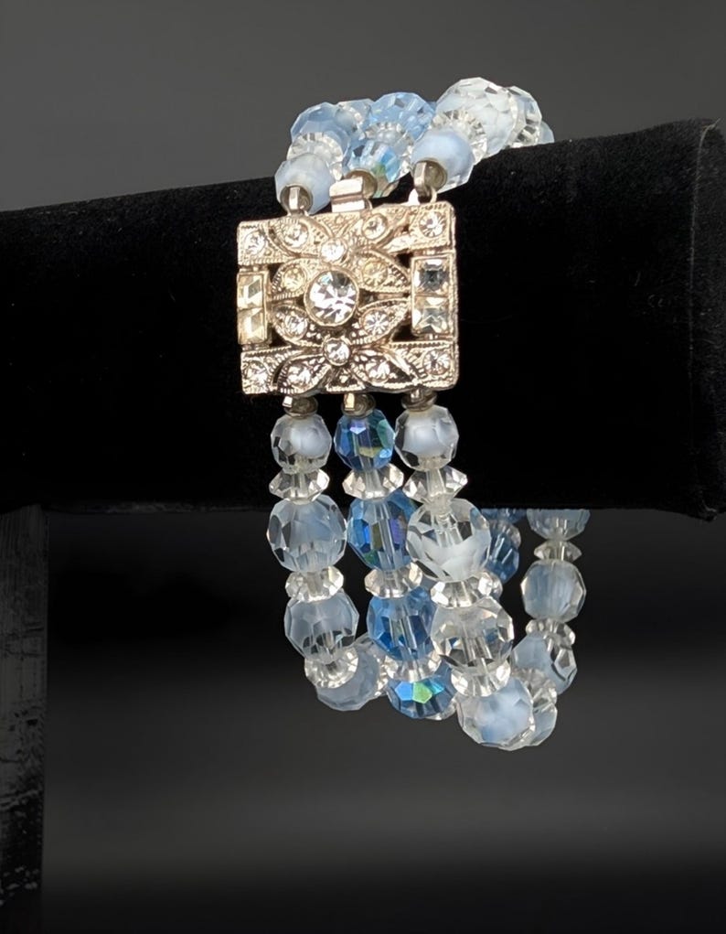May include: A vintage bracelet featuring three strands of light blue and clear crystal beads. The bracelet has a silver-tone square clasp adorned with clear rhinestones and intricate floral designs. The beads are faceted, reflecting light.