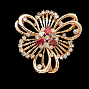 May include: A gold-toned floral brooch with a central cluster of red and clear gemstones. The brooch features a fan-like design with clear crystals and a curved, openwork frame. The background is solid black.