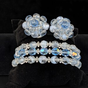 May include: A set of vintage jewelry featuring a pair of clip-on earrings and a bracelet. The earrings and bracelet are made of clear and light blue faceted beads. The jewelry is displayed on a black velvet surface.