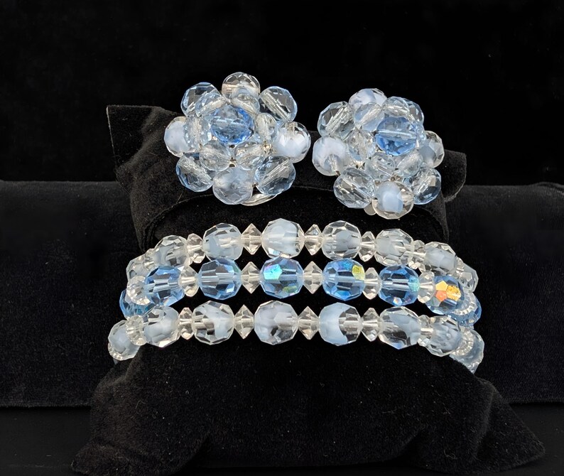 May include: A set of vintage jewelry featuring a pair of clip-on earrings and a bracelet. The earrings and bracelet are made of clear and light blue faceted beads. The jewelry is displayed on a black velvet surface.
