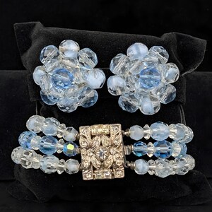 May include: A vintage jewelry set featuring a bracelet and matching earrings. The bracelet has three strands of clear and light blue beads with a decorative silver clasp. The earrings are clusters of similar beads. The set is displayed on a black velvet surface.